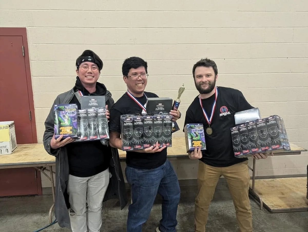 Three tournament winners standing together with medals around their necks, holding boxed HeroClix prizes.